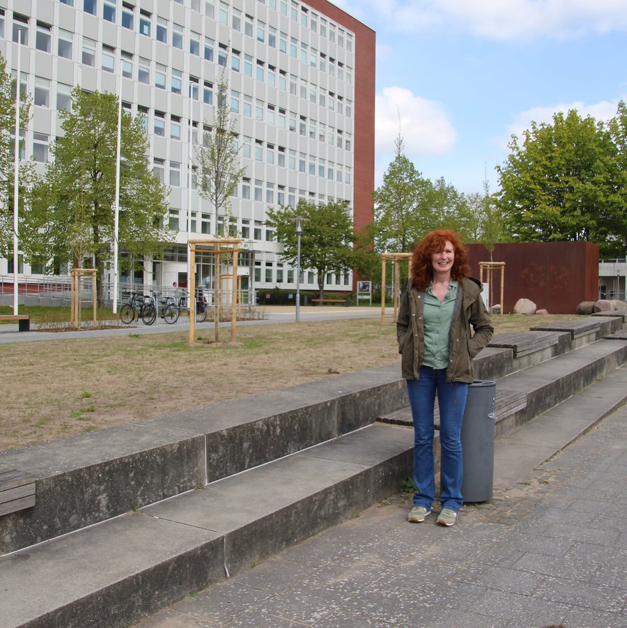 Eine Frau auf einem Platz vor zwei Treppenstufen. Im Hintergrund sind Bäume und ein Gebäude zu sehen