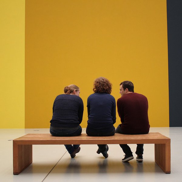 Drei Menschen sitzen auf einer Bank im Museum. Sie schauen auf eine gelbe Wand.