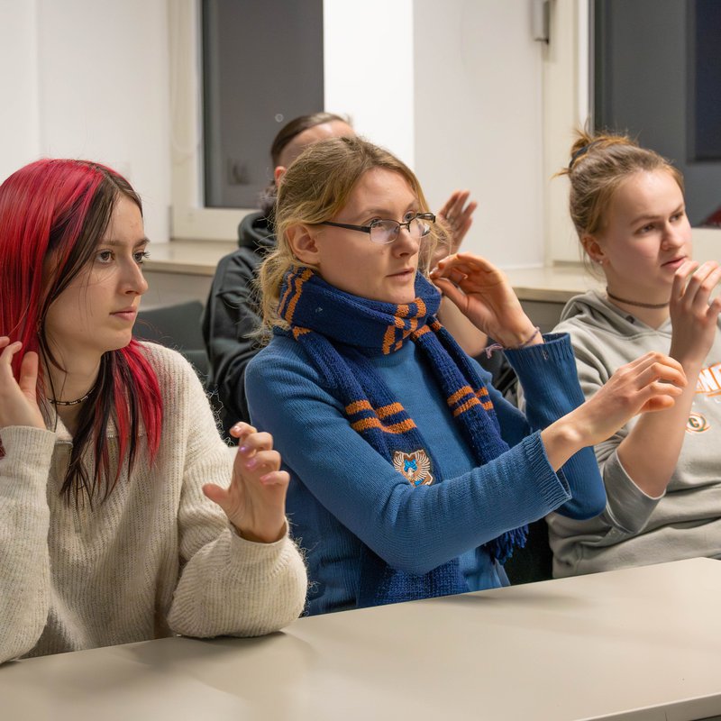 Drei Studentinnen sitzen in einer Reihe an einem Tisch. Sie halten ihre Hände in die Luft.