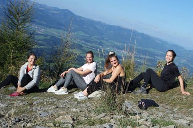 Jana und ihre Freundinnen sind zu sehen auf der Bergspitze, die Sonne scheint hell. Es wird eine Pause bei Schokolade und Snacks gemacht