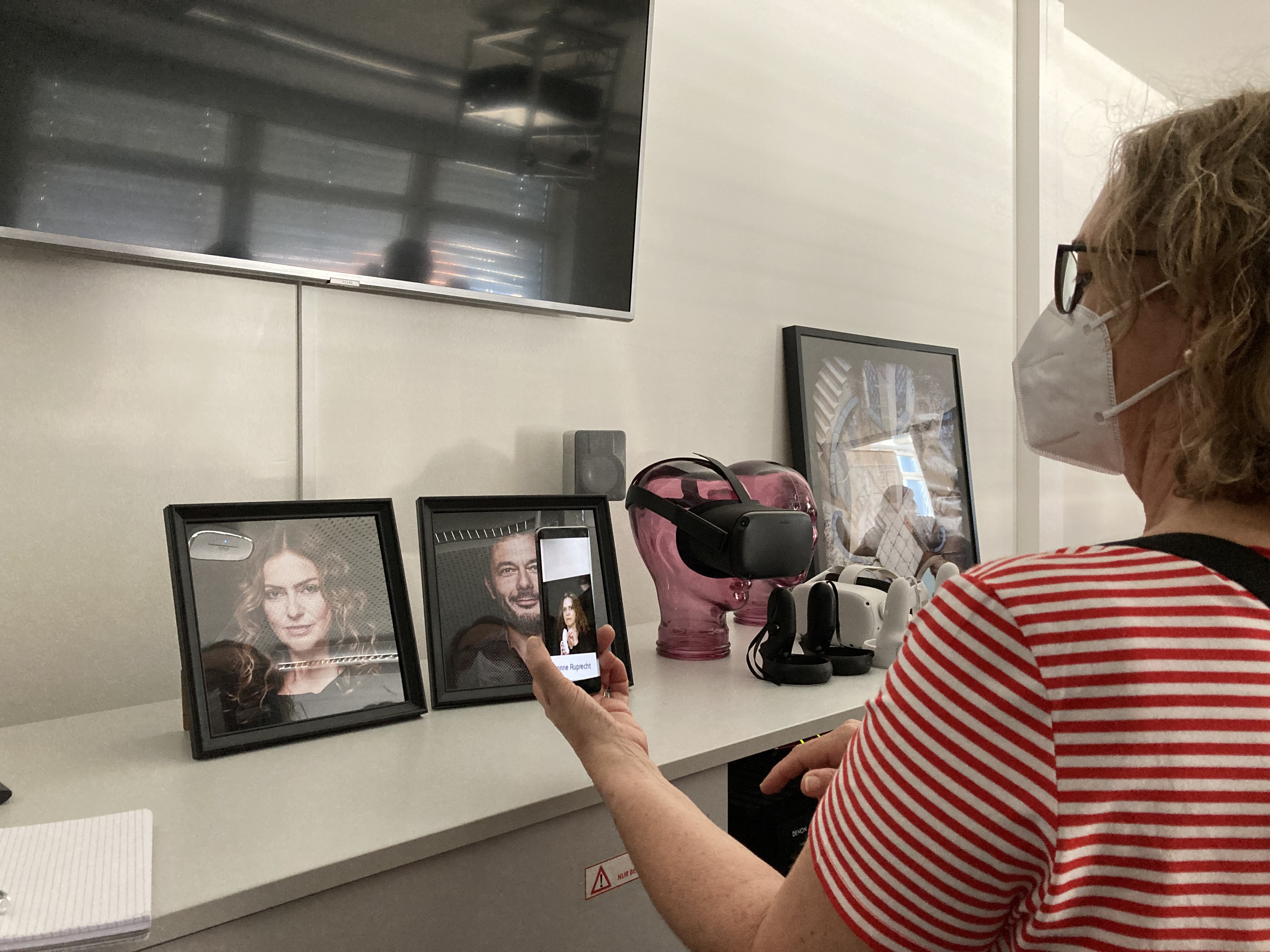 Eine Frau schaut auf ein Smartphone, auf dem ein Foto ein Video einer Frau zu sehen ist. Im Hintergrund ist die Frau aus dem Video auf einem Foto zu sehen.