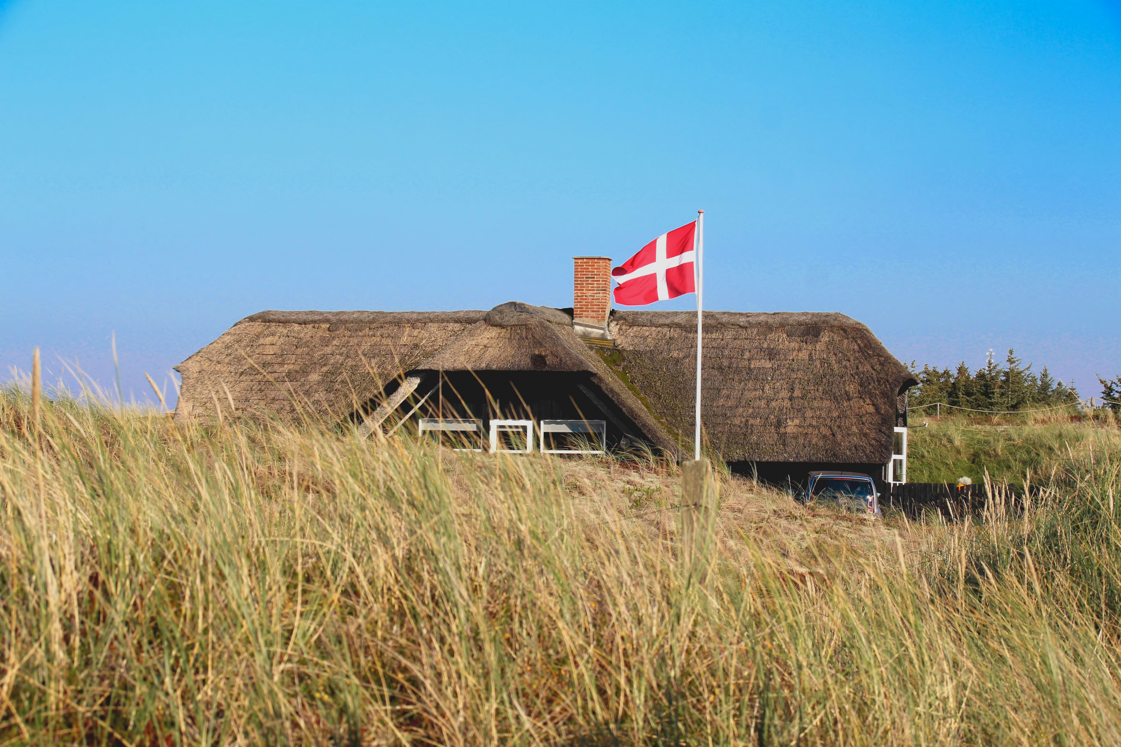 Dänemark Tourismus Ferienhaus in den Dünen ein Reetdachhaus hinter Dünen, davor weht die Nationalflagge Dänemarks