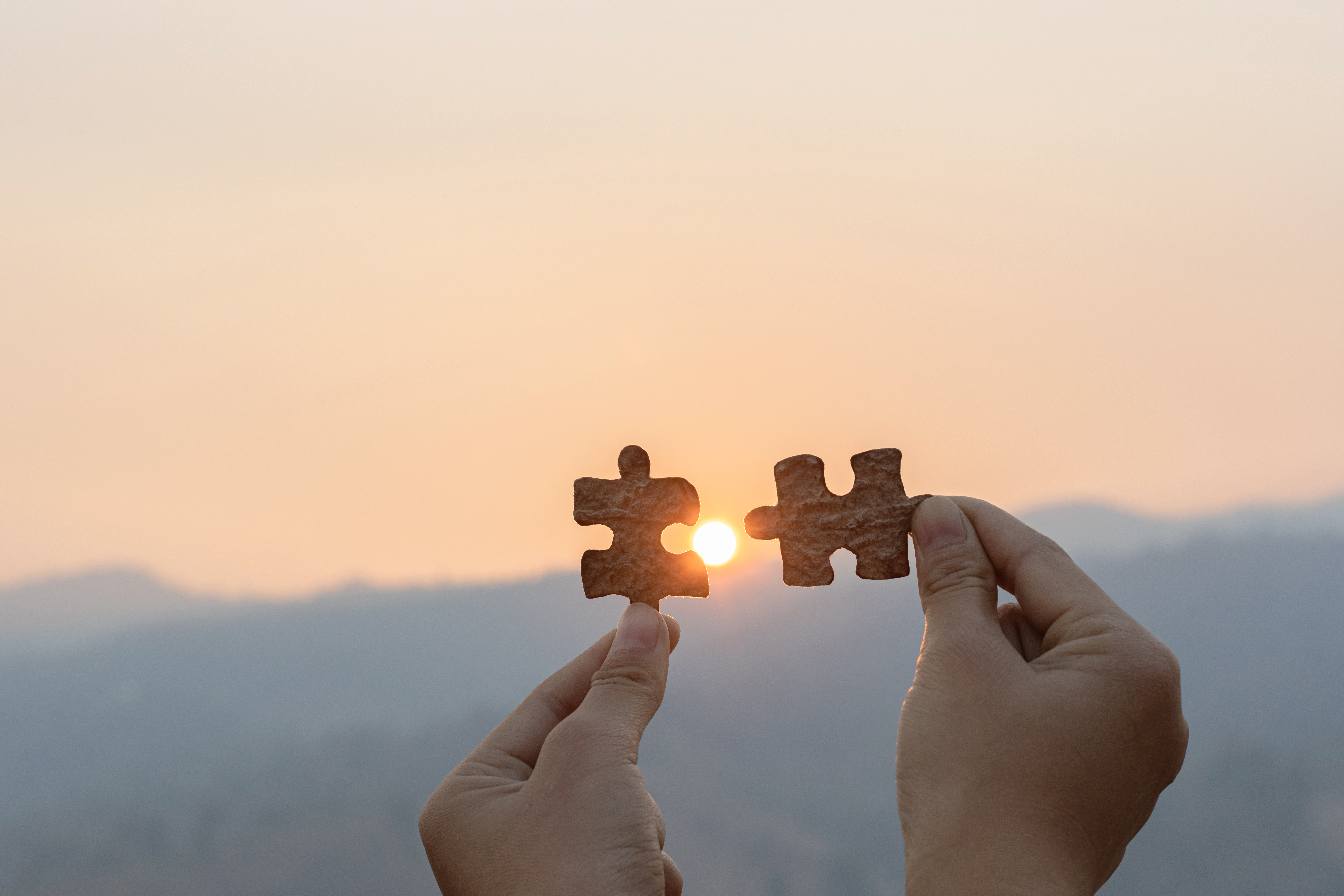 Closeup hand connecting jigsaw puzzle with sunset and mountains zwei Hände halten je ein Puzzleteil in die Sonne