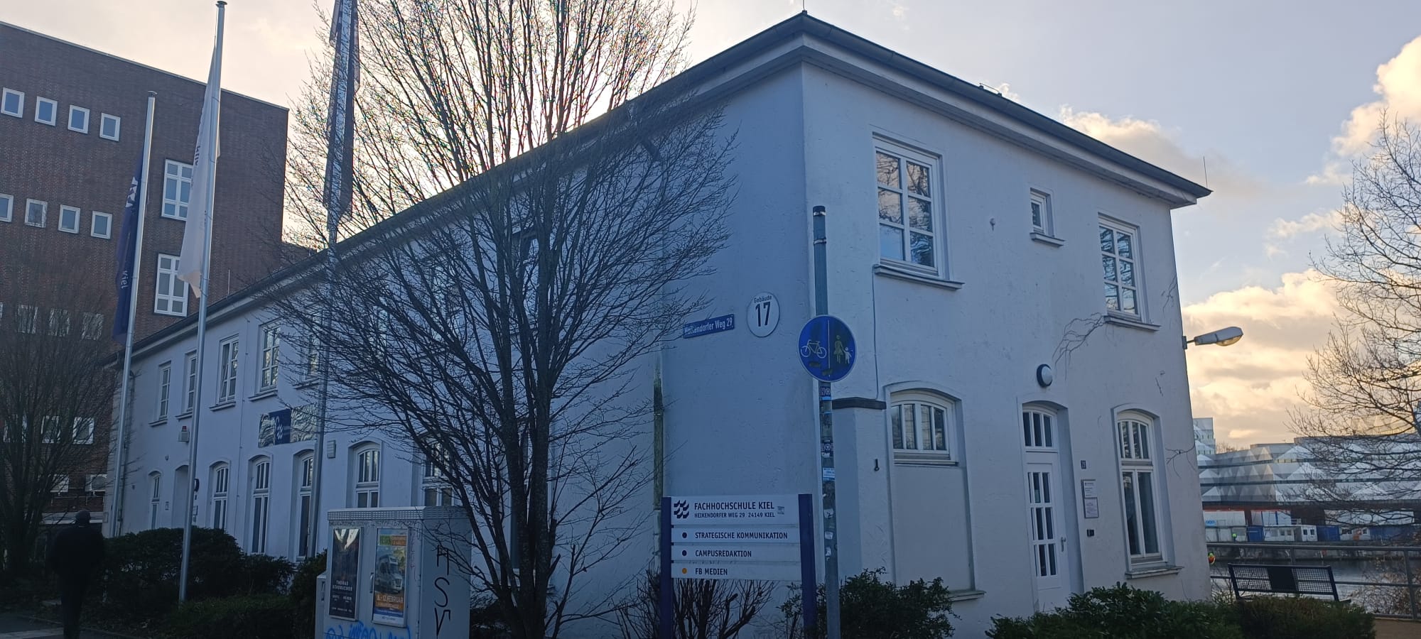 Ein weißes Gebäude mit zwei Fahnenmasten und einem kahlen Strauch davor.