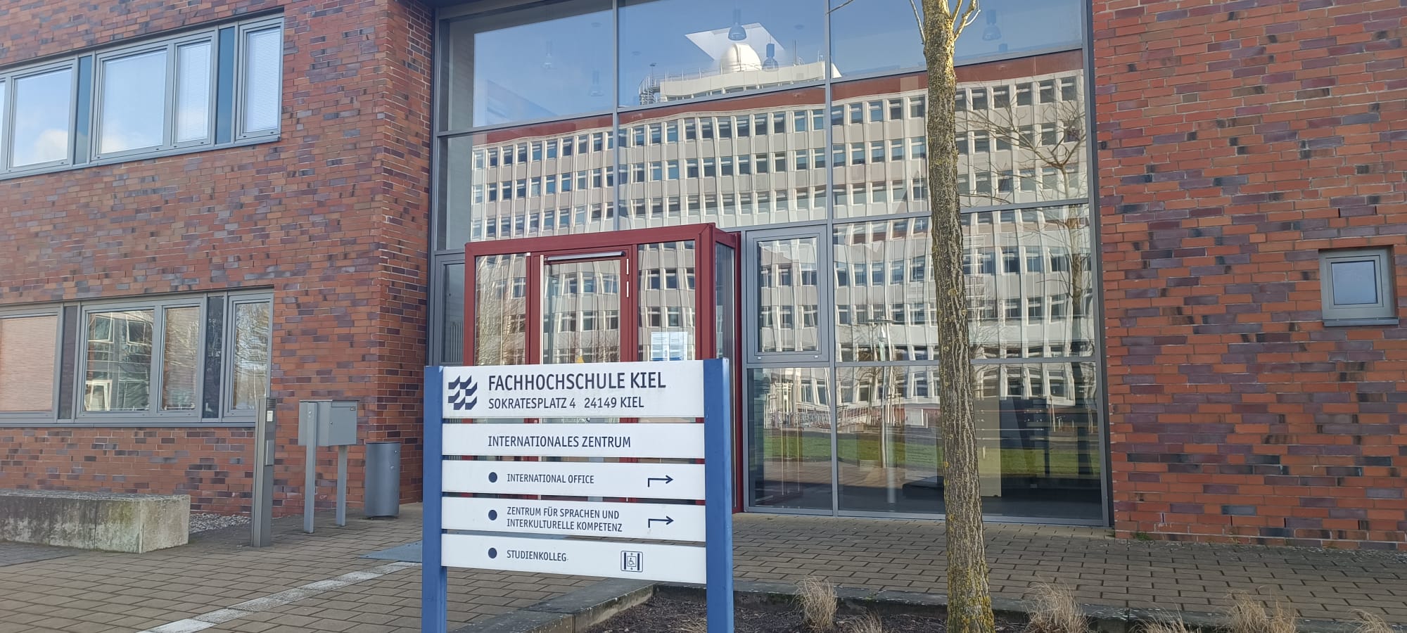 Ein rotbrauner Backstein-Bau mit großer Glasfassade, in welcher sich ein großes Gebäude spiegelt. Im Vordergrund steht ein Schild, welches das International Office ausweist.