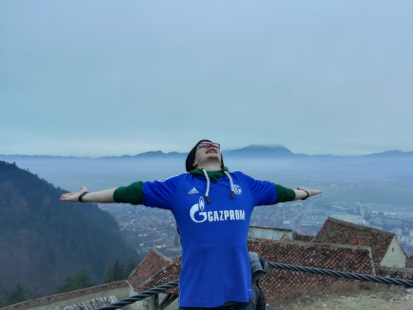 Ein Mann in einem Schalke 04 Trikot posiert mit weit geöffneten Armen auf einem Dach. Im Hintergrund ist eine von Bergen umgebene und in Nebel getauchte Stadt.