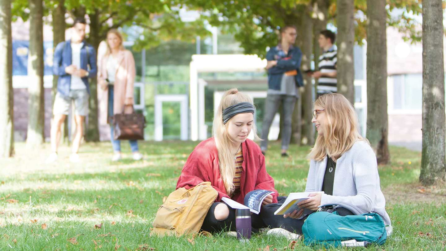 Zwei Studentinnen sitzen auf einer Wiese und lesen in Infobroschüren.