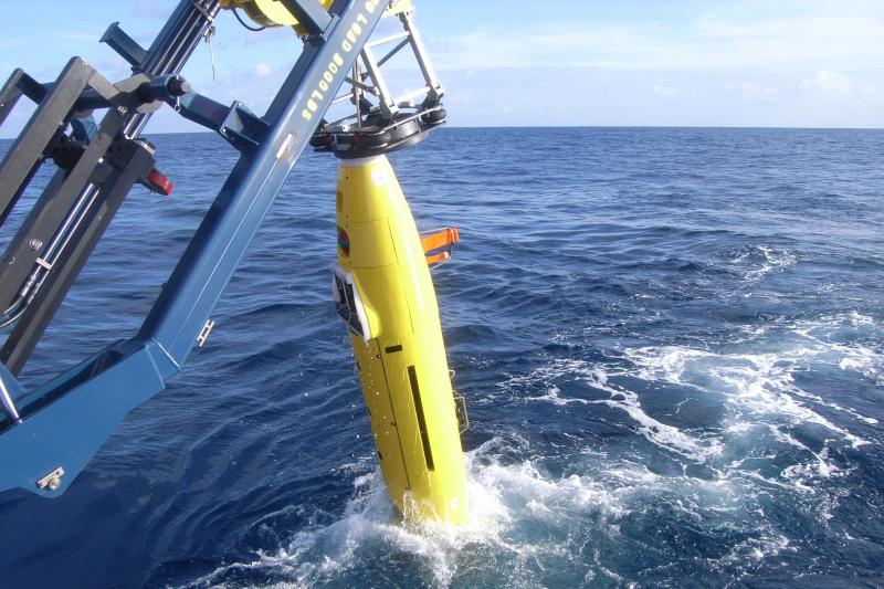 Mitten im Meer wird ein gelbes torpedoartiges Geraät, mit einem Kran, aus dem Wasser geholt.