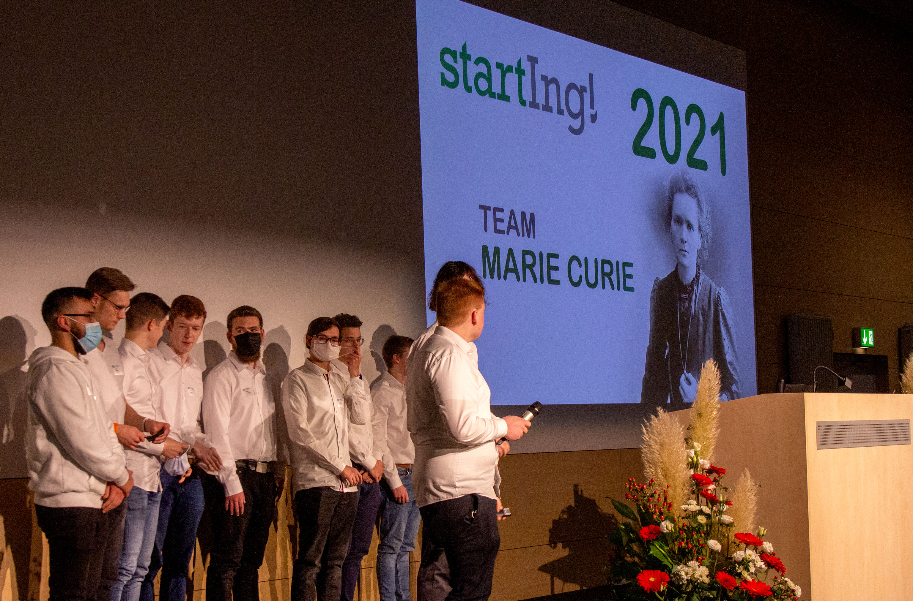 Eine Gruppe Männer in weißen Hemden auf der Bühne, im Hintergrund das Titelbild ihrer Präsentation: Team Marie Curie