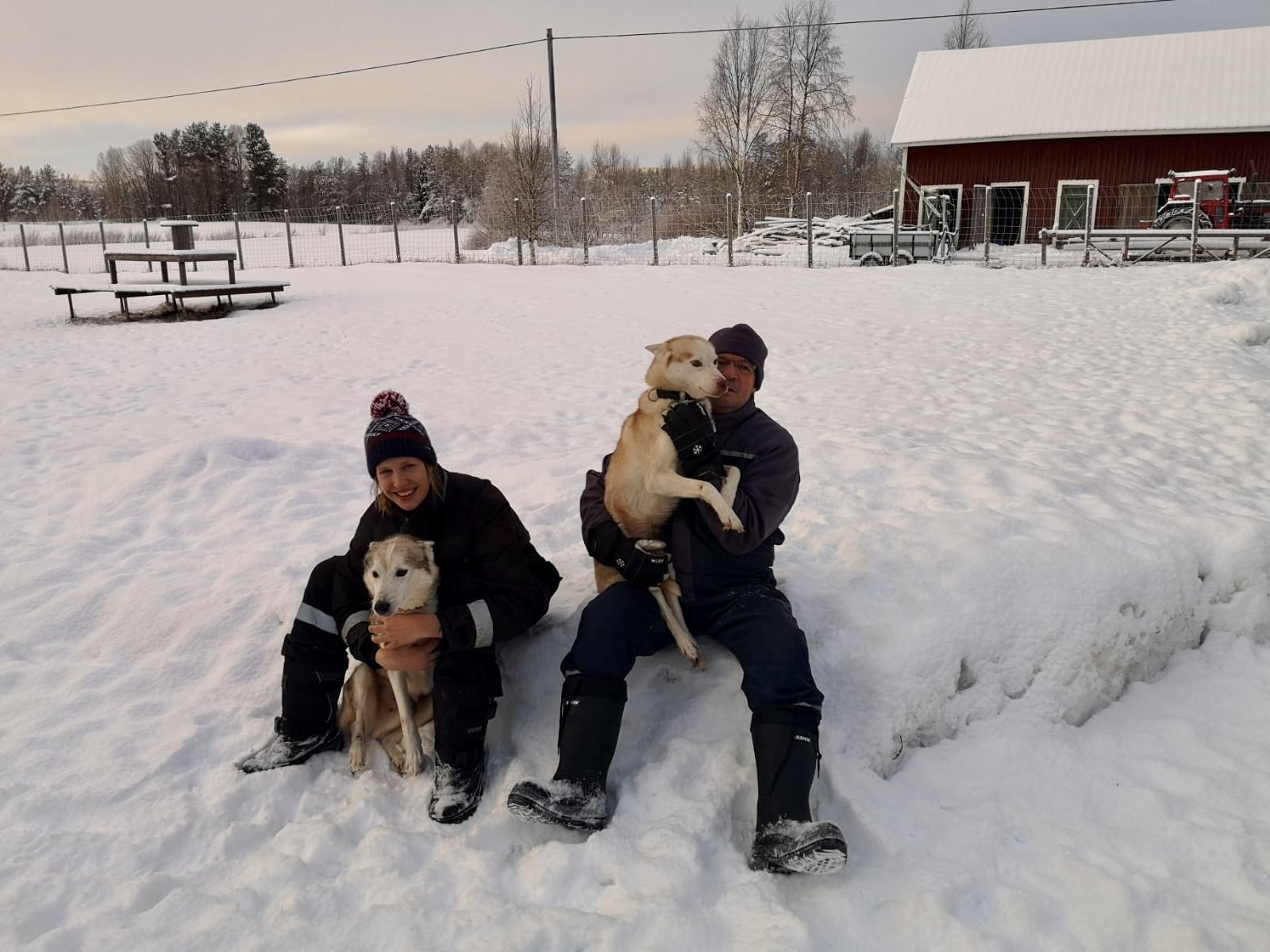 Eine Frau und ein Mann sitzen im Schnee und halten jeweils einen Hund im Arm. Sie haben beide einen Schneeanzug an und eine Mütze auf.