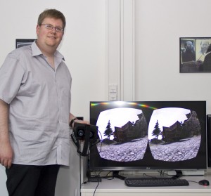Ein Mann in grauem Kurzarmhemd, steht neben einem Monitor.
