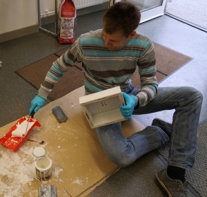 Ein Mann sitzt auf einer Werkplatte und streicht einen Rahmen.