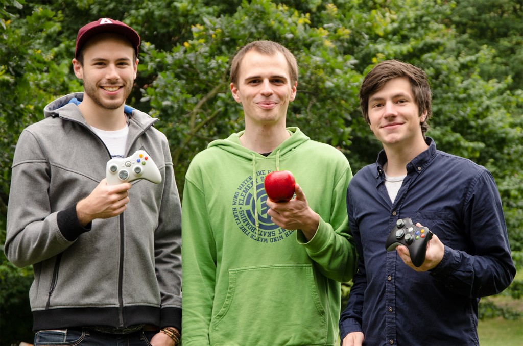 Drei Männer stehen nebeneinander im Freien. Zwei von ihnen halten Controller und einer einen roten Apfel in der Hand.