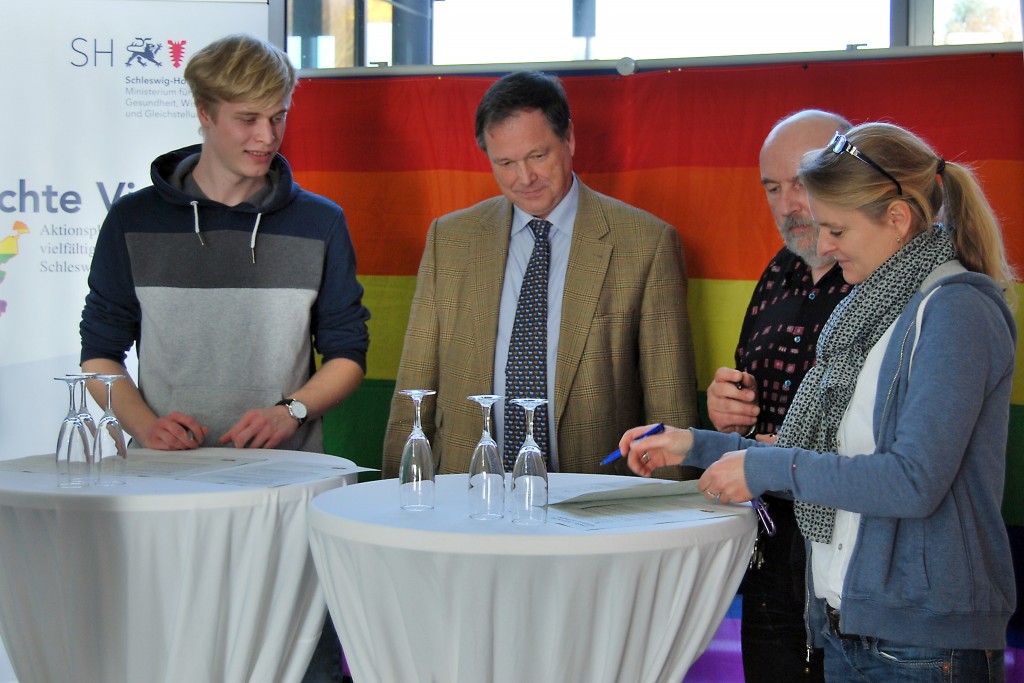 Zwei Männer und zwei Frauen stehen an einem Tisch und unterschreiben ein Schriftstück.