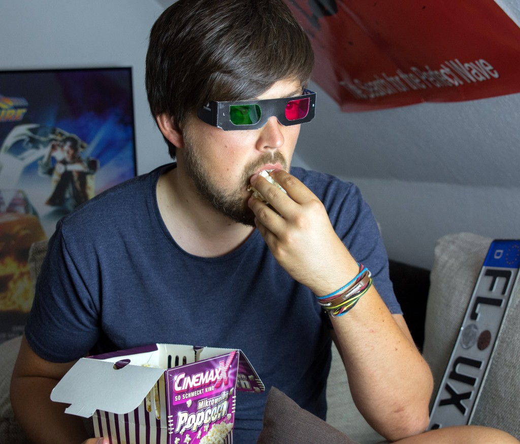 Ein Mann mit 3D-Brille sitzt in seinem Zimmer und isst Popcorn.