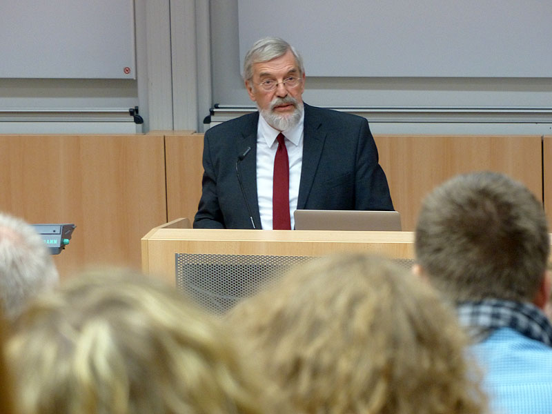 Ein Mann mit roter Krawatte steht am Rednerpult eines Hörsaals und blickt auf seine Zuhörenden.