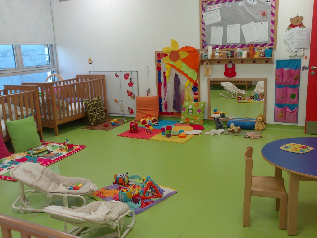 Ein Raum in einer Krippe ist mit grünem Boden ausgelegt. Es befinden sich viele Spielzeuge und Betten in dem Raum.