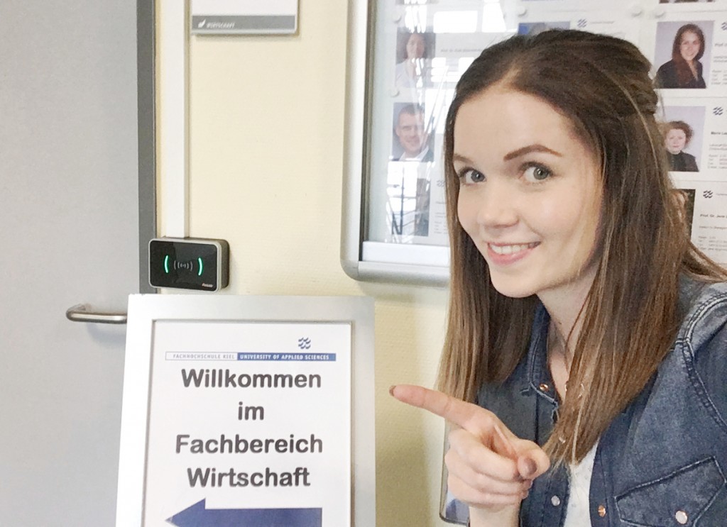 Eine Studentin zeigt mit dem Finger auf das Willkommensschild des Fachbereichs Wirtschaft der FH Kiel.