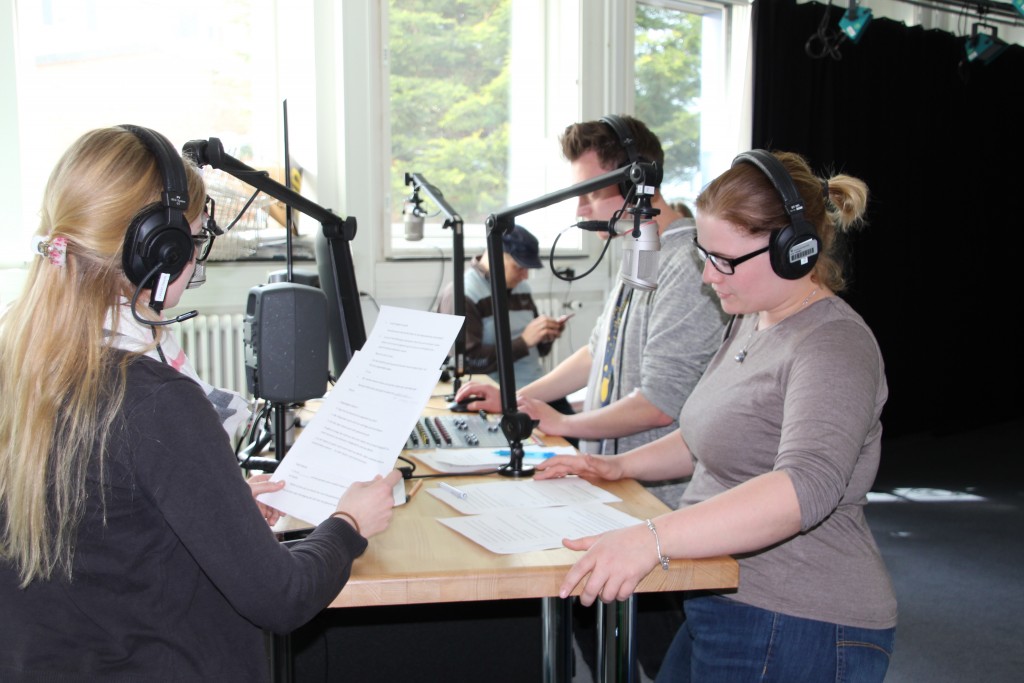 Zwei Frauen und ein Mann stehen mit Kopfhörern ausgestattet vor ihren Mikrofonen.