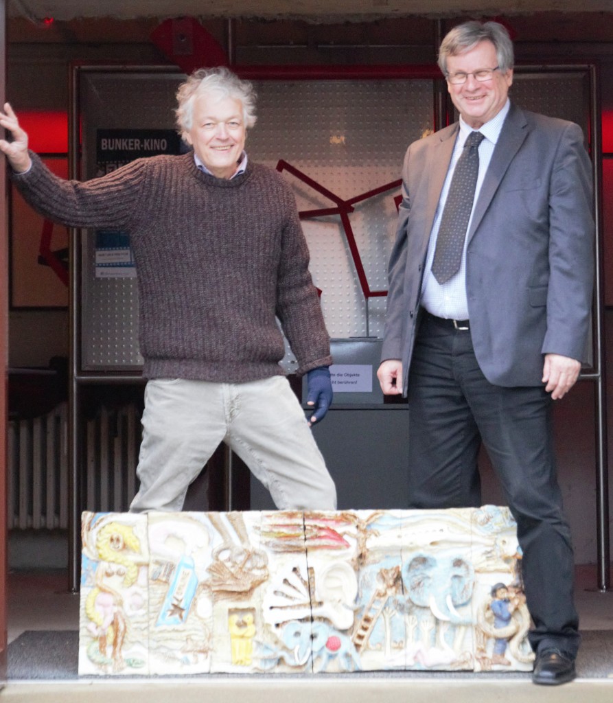 Zwei Männer, einer in Anzug, der ander in Strickpullover und Cordhose stehen auf einer Bühne und präsentieren Kunst.