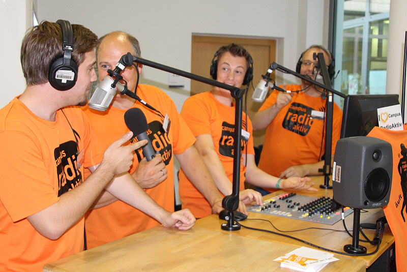 Vier Männer in orangenen Shirts sprechen angeregt vor zwei Mikrofonen.