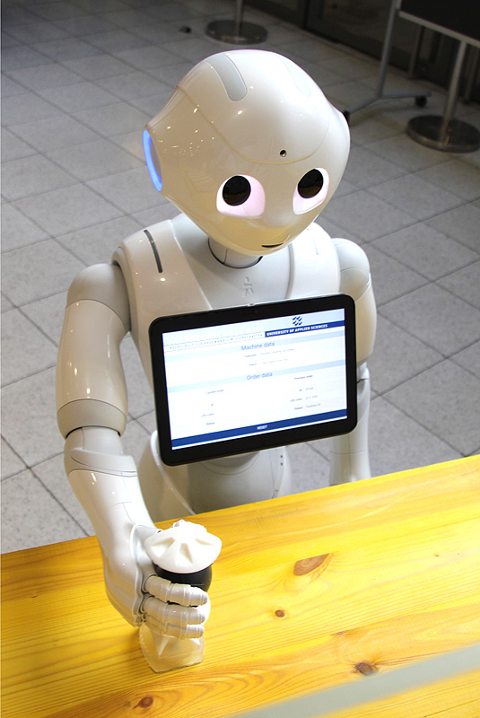 Ein weißer Roboter mit Tablet auf der Brust montiert stellt etwas auf einem Tisch ab.