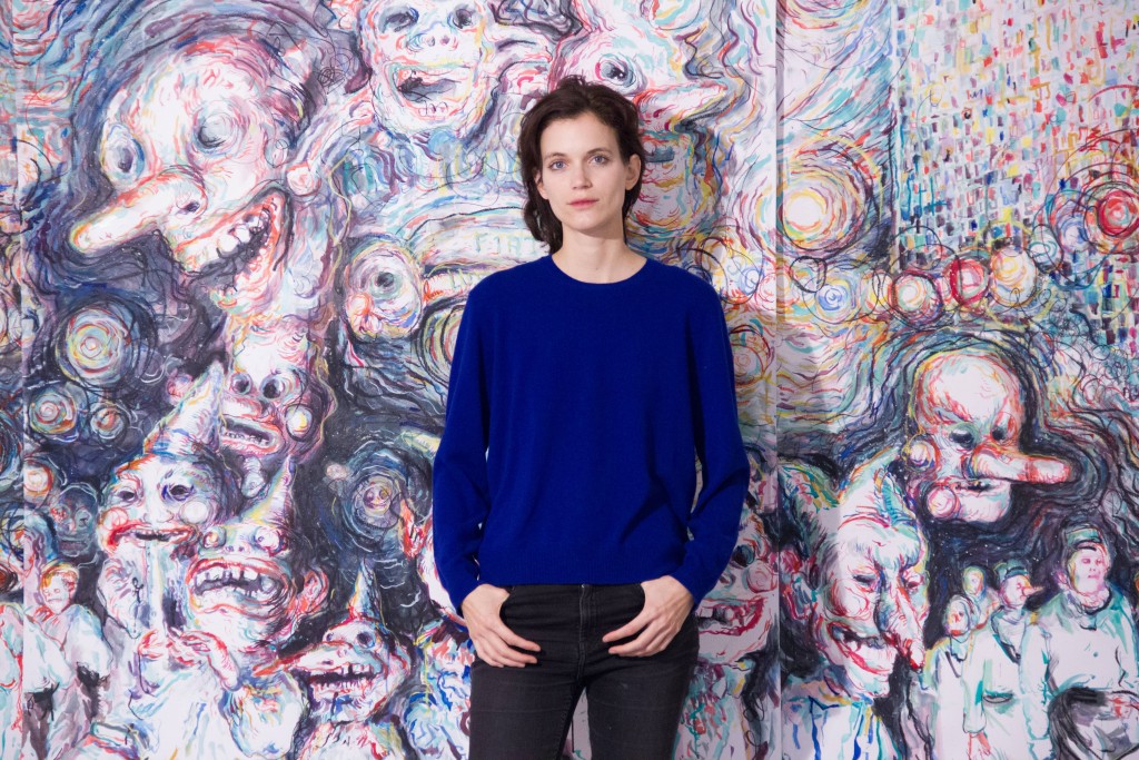 Eine Frau in blauem Pullover posiert vor einem bizarren Gemälde.