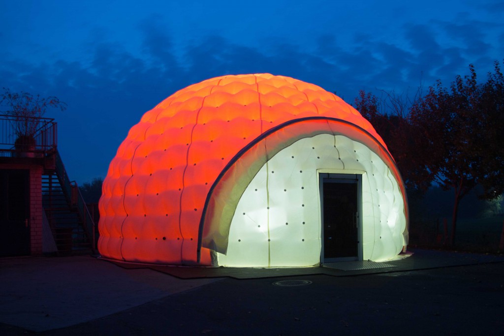 Ein großes Zelt in der Form eines Iglu, ist mit einer orangenen Plane überdeckt und von innen stark beleuchtet.