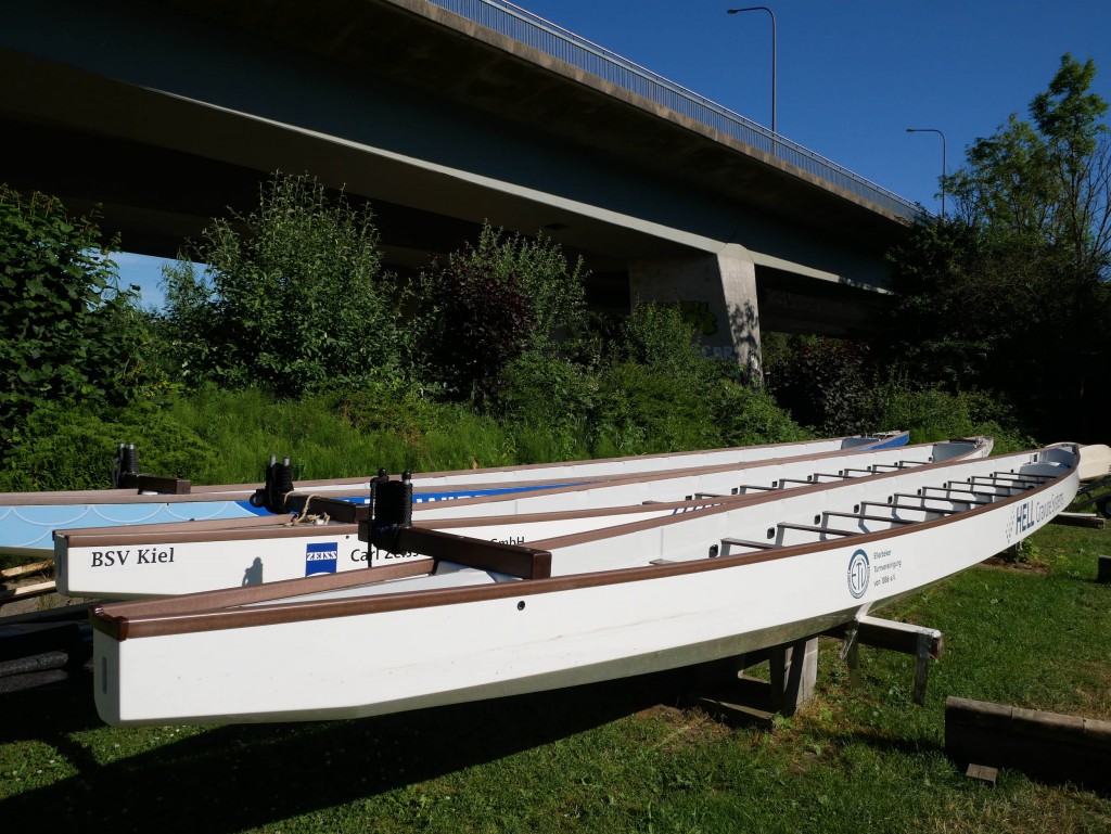 Drei Ruderboote stehen auf Böcken nebeneinander. Im Hintergrund ist eine Brücke zu sehen.