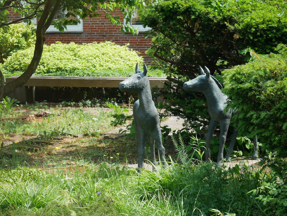 Zwei Pferdeskulpturen stehen auf einer Grünfläche, in der Nähe eines Buschs.