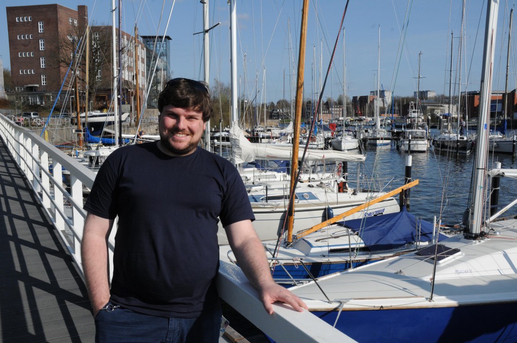 Ein Mann steht am Bootssteg, eines Yachthafens  und lächelt in die Kamera.