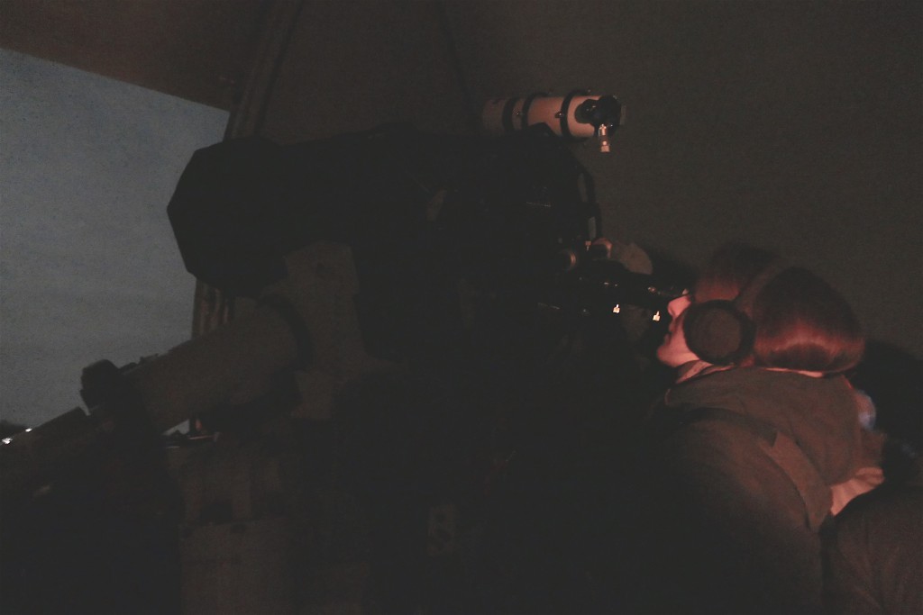 Eine FRau mit Ohrenschützern schaut durch ein Teleskop in den Abendhimmel hinaus.