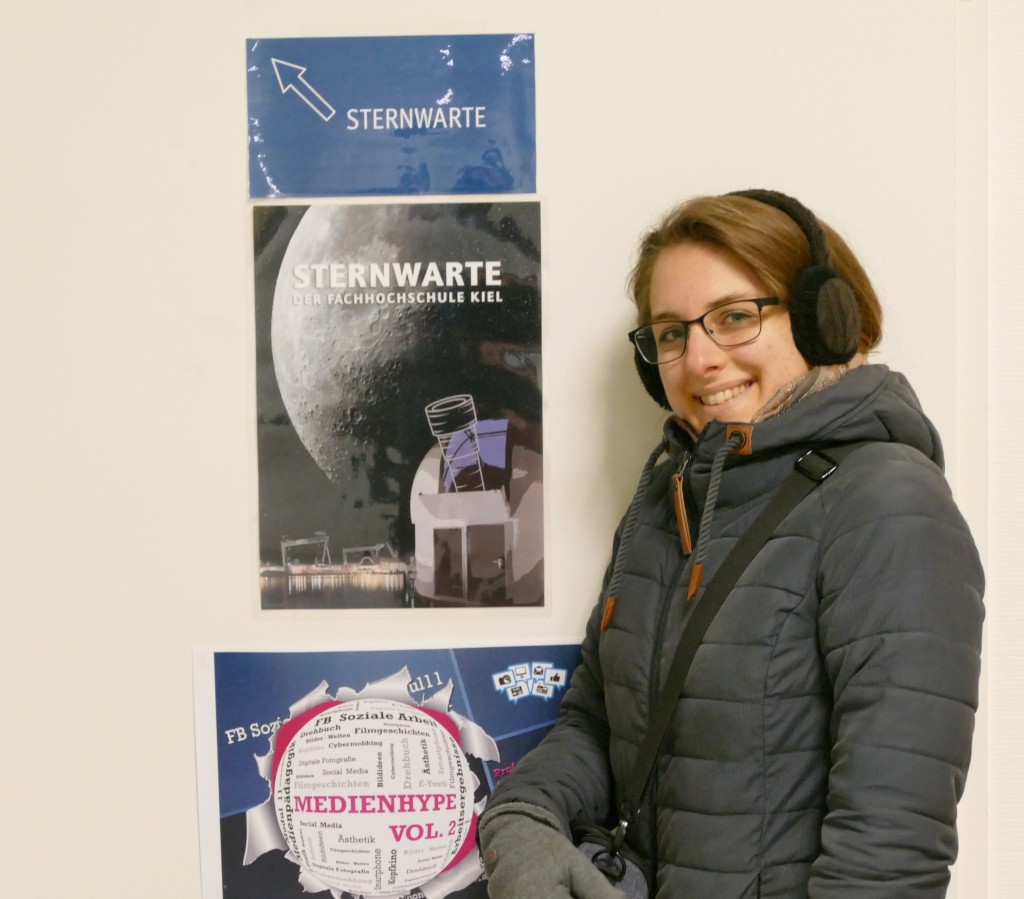 Eine Frau mit Winterjacke und Ohrenwärmern posiert vor Plakaten der Sternenwarte Fachhochschule Kiel.